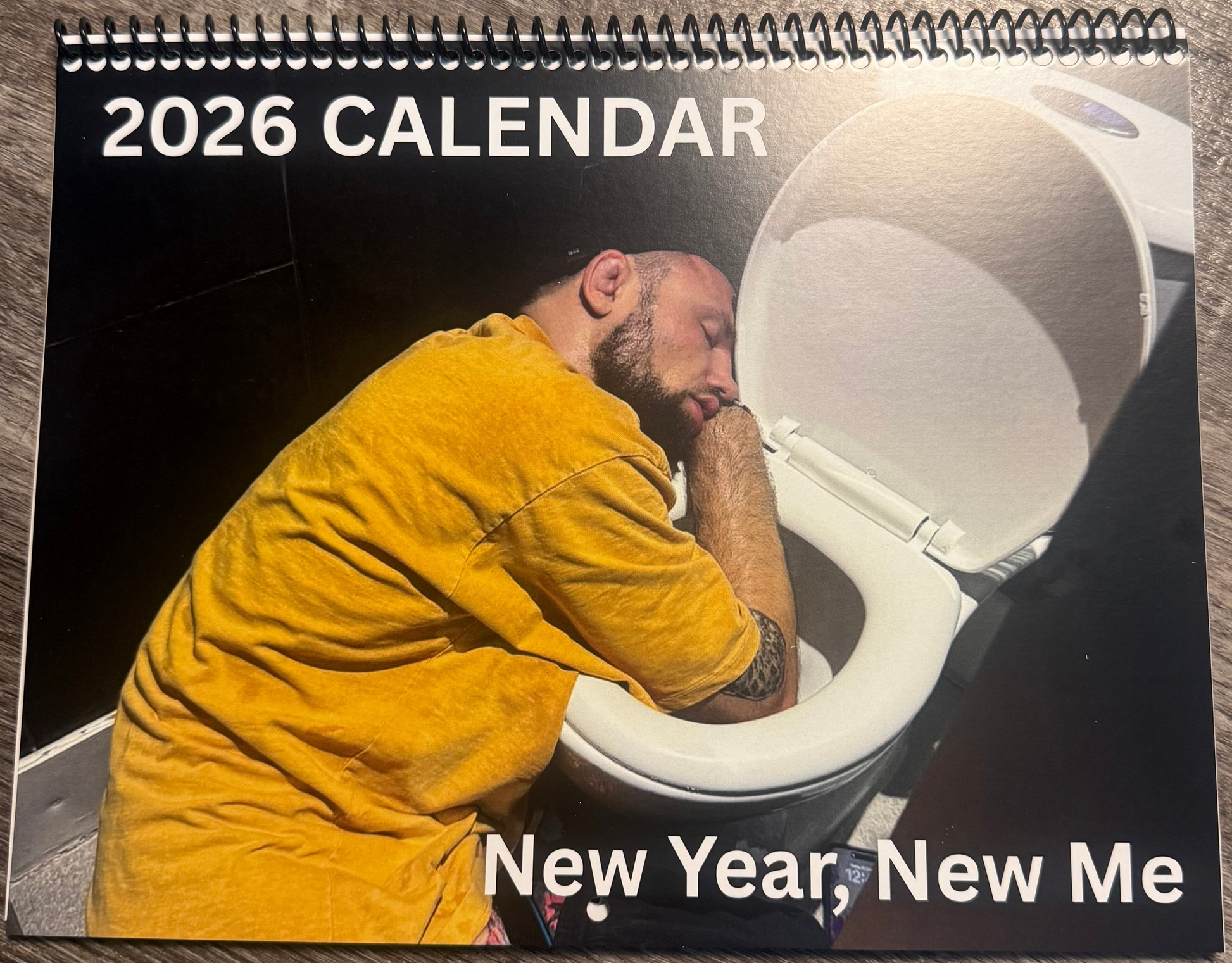 2026 calendar with a man in a yellow shirt sitting on a toilet, titled 'New Year, New Me', on a wooden surface.